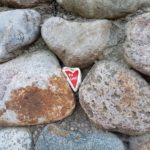 Ein herzförmiger Stein mit dem in Rot aufgemalten Wort „Love“ liegt eingebettet zwischen verschiedenen runden und unregelmäßig geformten Felsen mit natürlichen Braun- und Grautönen und gesprenkelten Mustern.