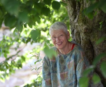 Eine ältere Frau mit kurzem weißem Haar und einer bunten Bluse lächelt herzlich und lugt durch die üppigen grünen Blätter eines Baumes in einer sonnigen Umgebung im Freien.